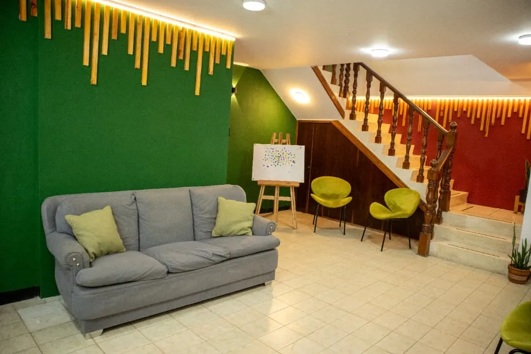La sala de Oikos: un sofá gris con cojines verdes, dos sillas verdes y la escalera de madera con luz cálida.
