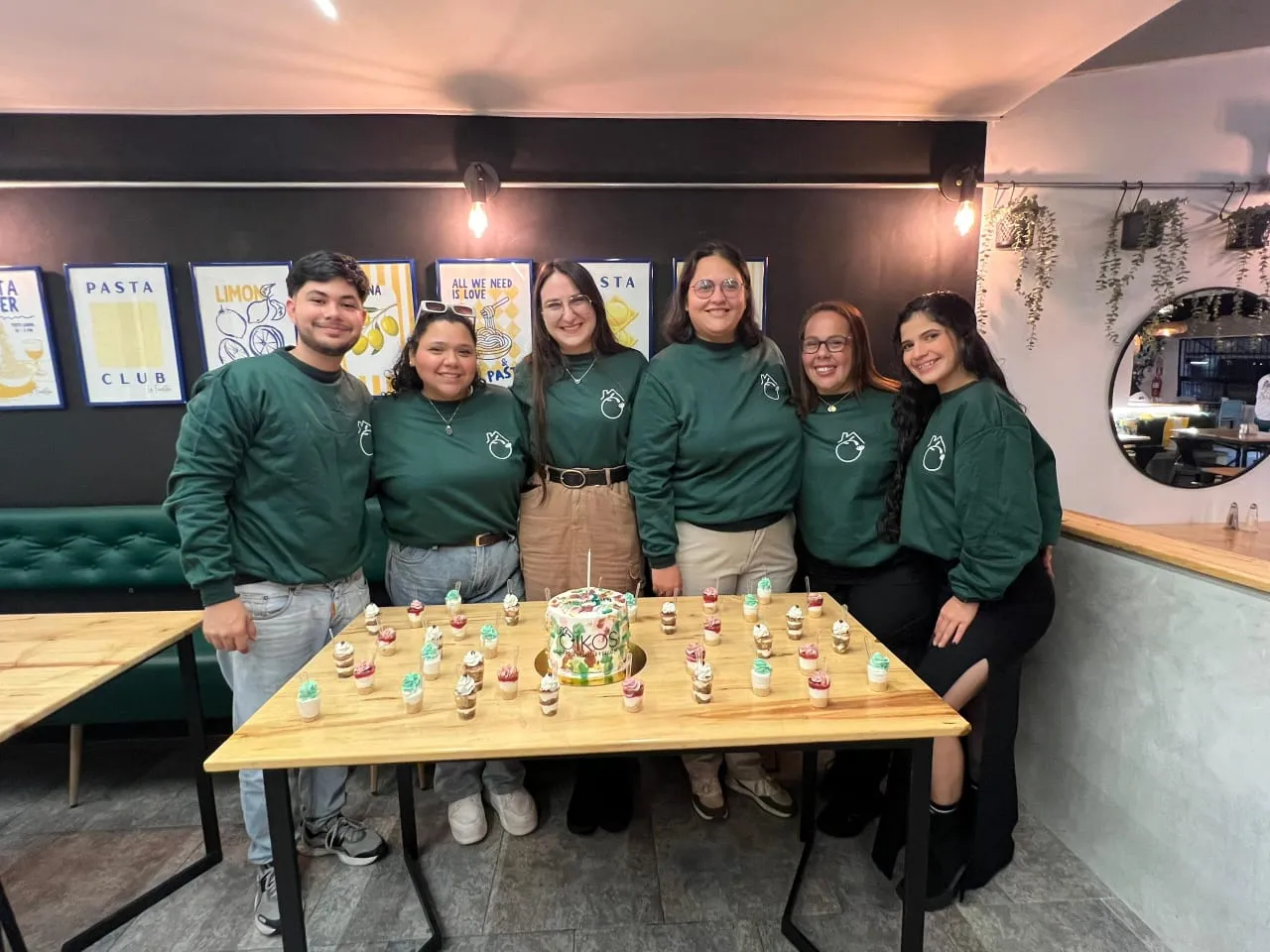 Seis personas del equipo de Oikos, con sudaderas verdes de la casa, alrededor de una torta con el logo de Oikos.